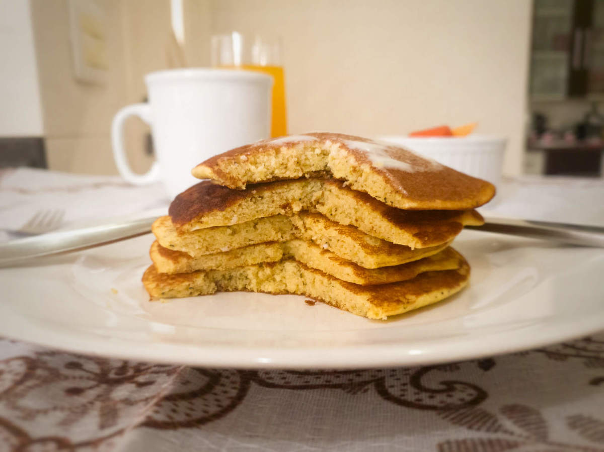 mesa com panquecas de fubá e requeijão cortadas no prato, caneca, talheres, ramequim com fruta e copo de suco de laranja