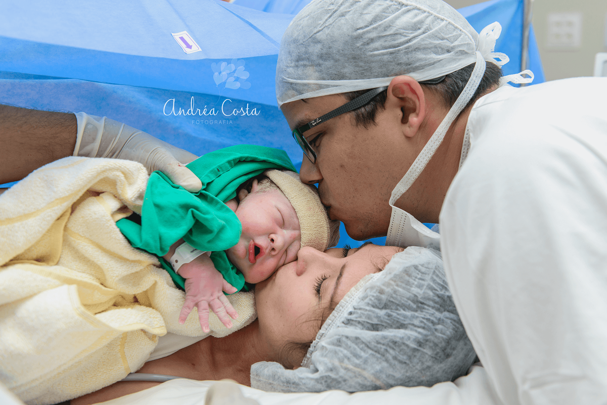 Pai e mãe beijando o filho recém nascido logo após o parto, por Andréa Costa