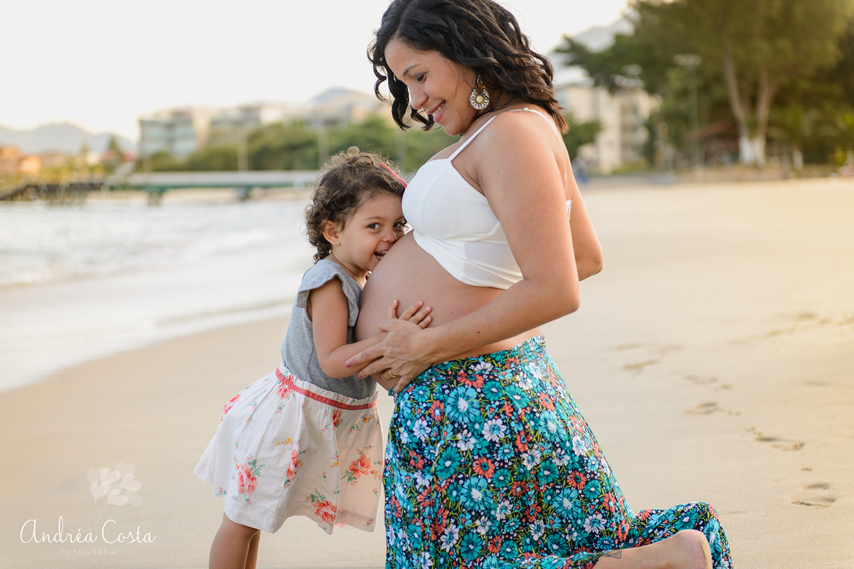 Irmãzinha beijando o irmão na barriga da mãe na praia, por Andréa Costa