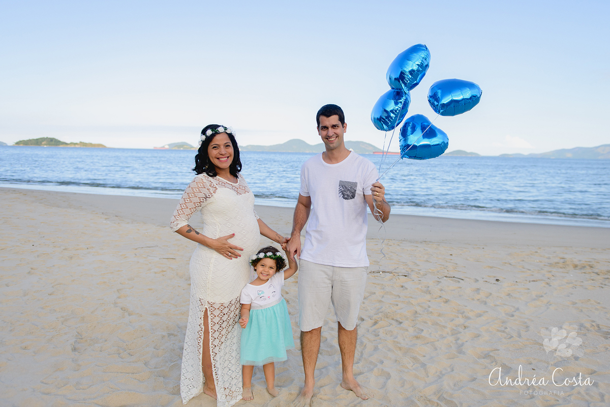 Grávida com vestido branco com a família segurando balões azuis na praia de Sahy, por Andréa Costa