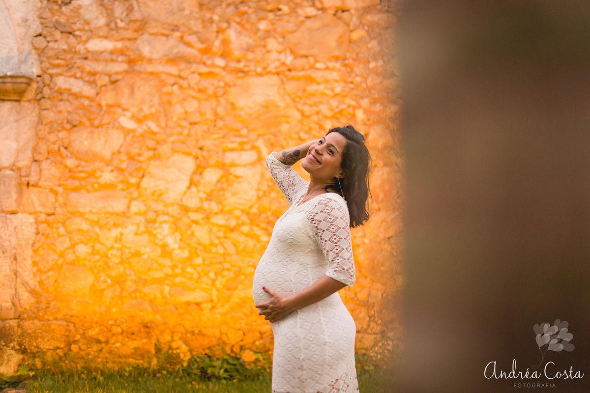 Mãe com a mão no cabelo na frente de uma parede de pedras , com luz amarela na praia de Sahy, RJ, por Andréa Costa