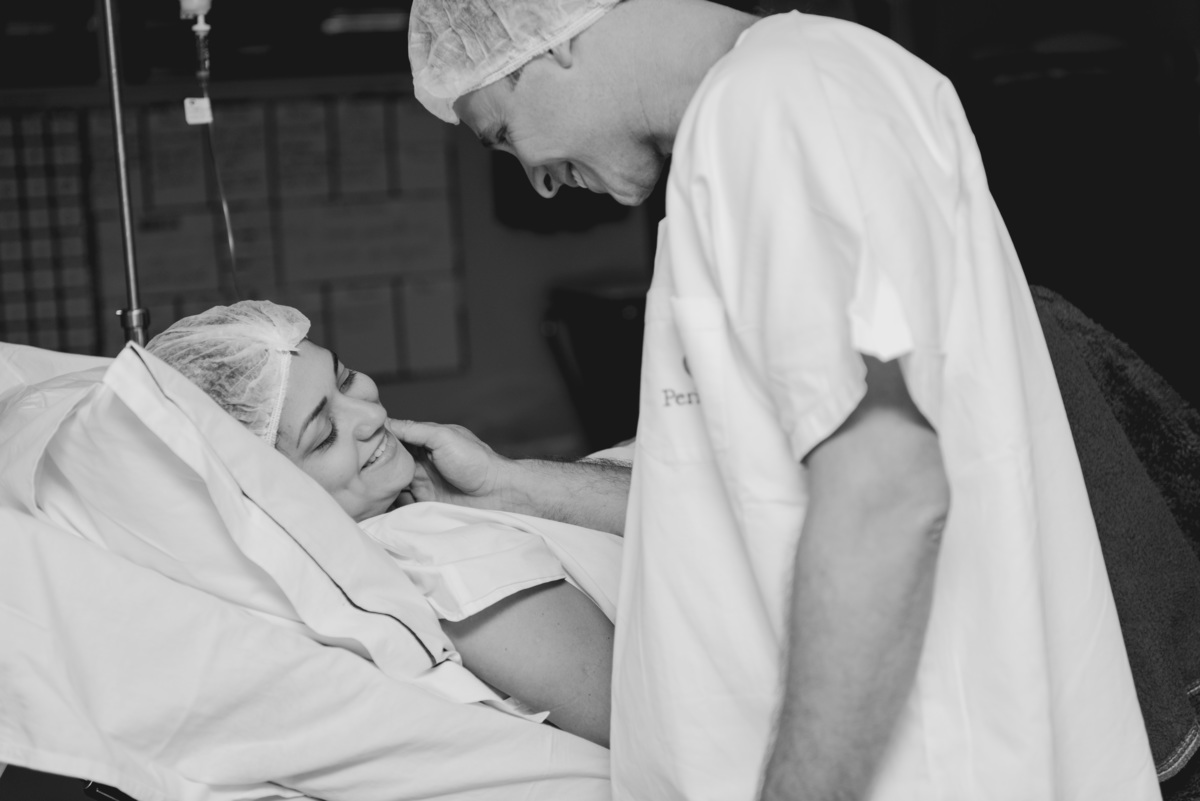 Grávida sorrindo para o marido dentro do centro cirúrgico da perinatal no rio de janeiro, por Andréa Costa