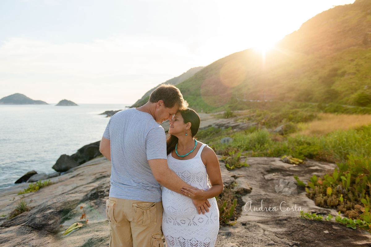 Casal se olhando com sol no fundo em cima das pedras na praia da Barra da Tijuca no Rio de Janeiro, por Andréa Costa