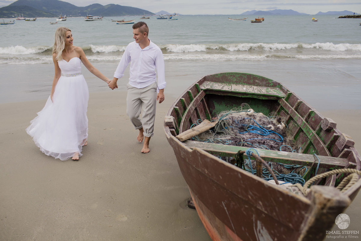 casal andando de mãos dadas ao lado de um barco na praia de garopaba sc