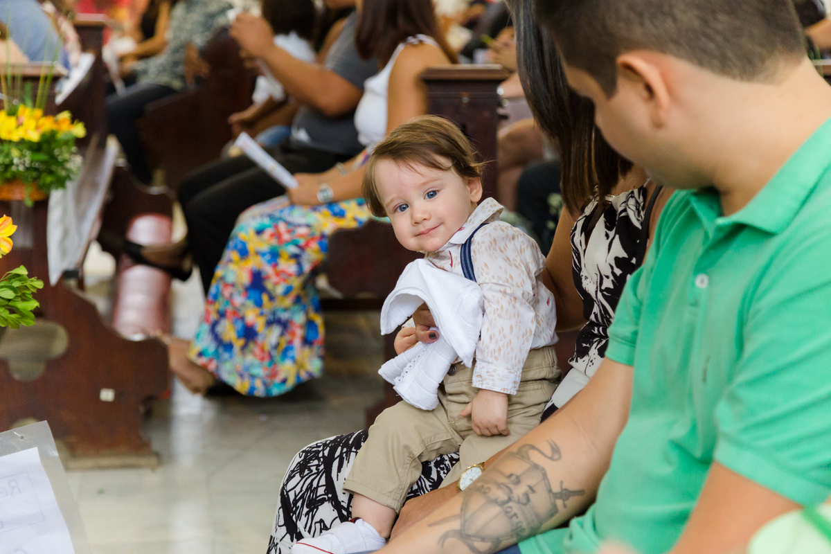Criança sorri para foto enquanto o padrinho o observa durante cerimônia de batismo
