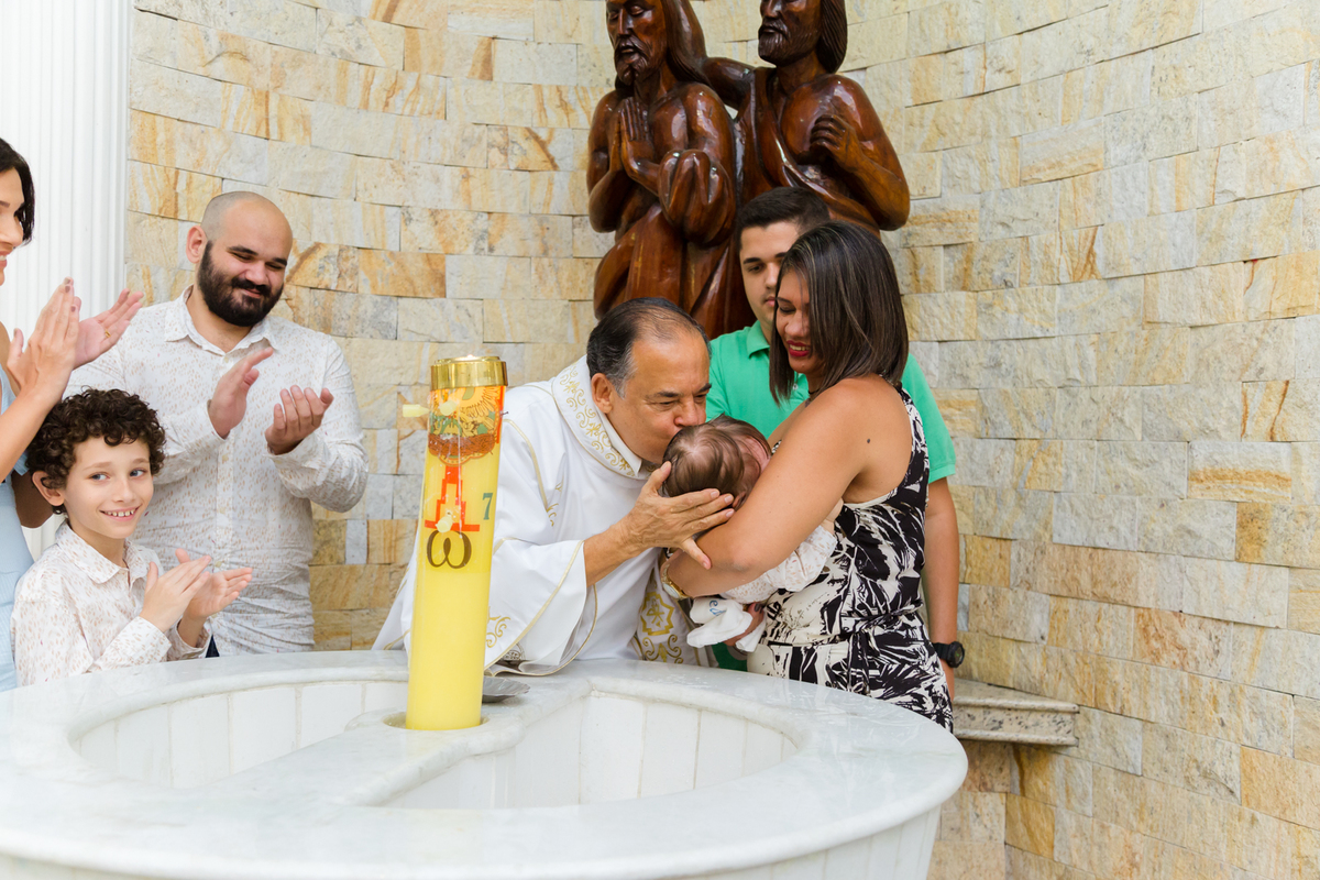 Na pia batismal padre beija criança após jogar água benta e faimilares aplaudem.