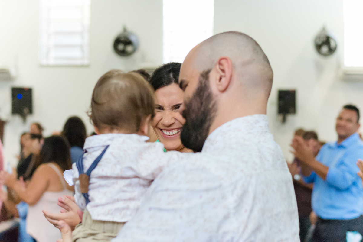 Mãe sorri para o filho enquanto o pai brinca com ele durante a cerimônia de batismo