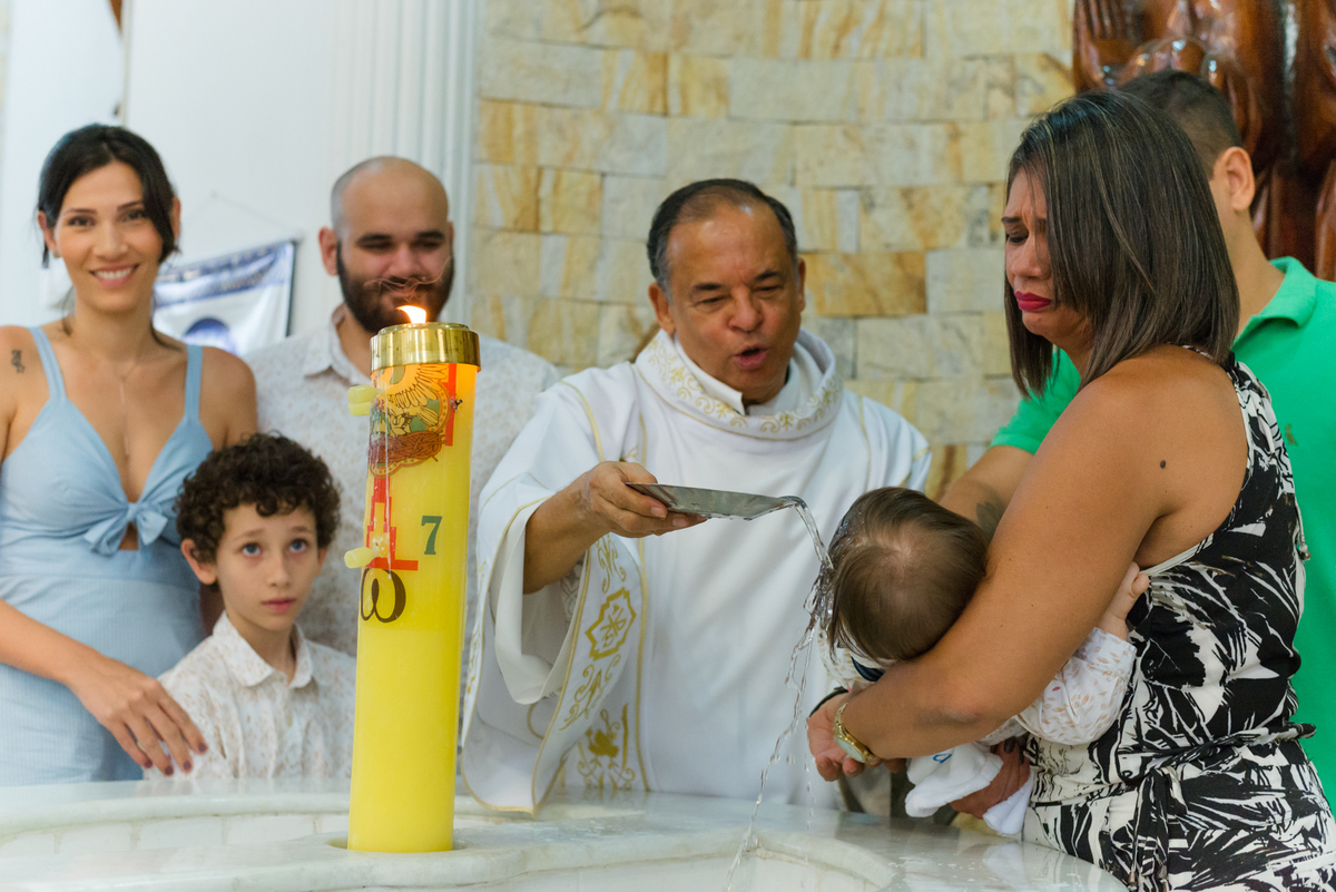 Padre derrama água benta sobre a cabeça da criança na pia batismal enquanto a madrinha o segura e os pais assistem