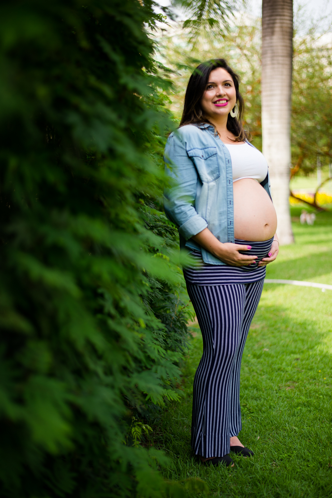 Mamãe feliz com as mãos no seu barrigão registrado pelo fotógrafo de gestante Nós Dois Fotografia na Barra da Tijuca RJ num belo jardim de fundo