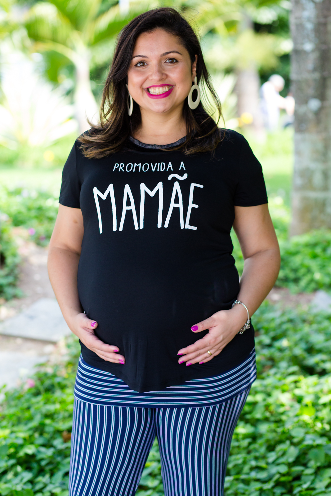 Mamãe feliz com as mãos no seu barrigão registrado pelo fotógrafo de gestante Nós Dois Fotografia na Barra da Tijuca RJ num belo jardim de fundo
