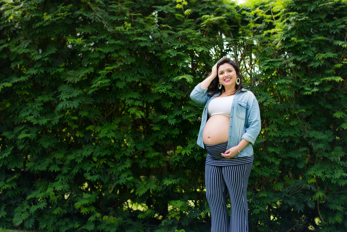 Mamãe feliz com as mãos no seu barrigão registrado pelo fotógrafo de gestante Nós Dois Fotografia na Barra da Tijuca RJ num belo jardim de fundo