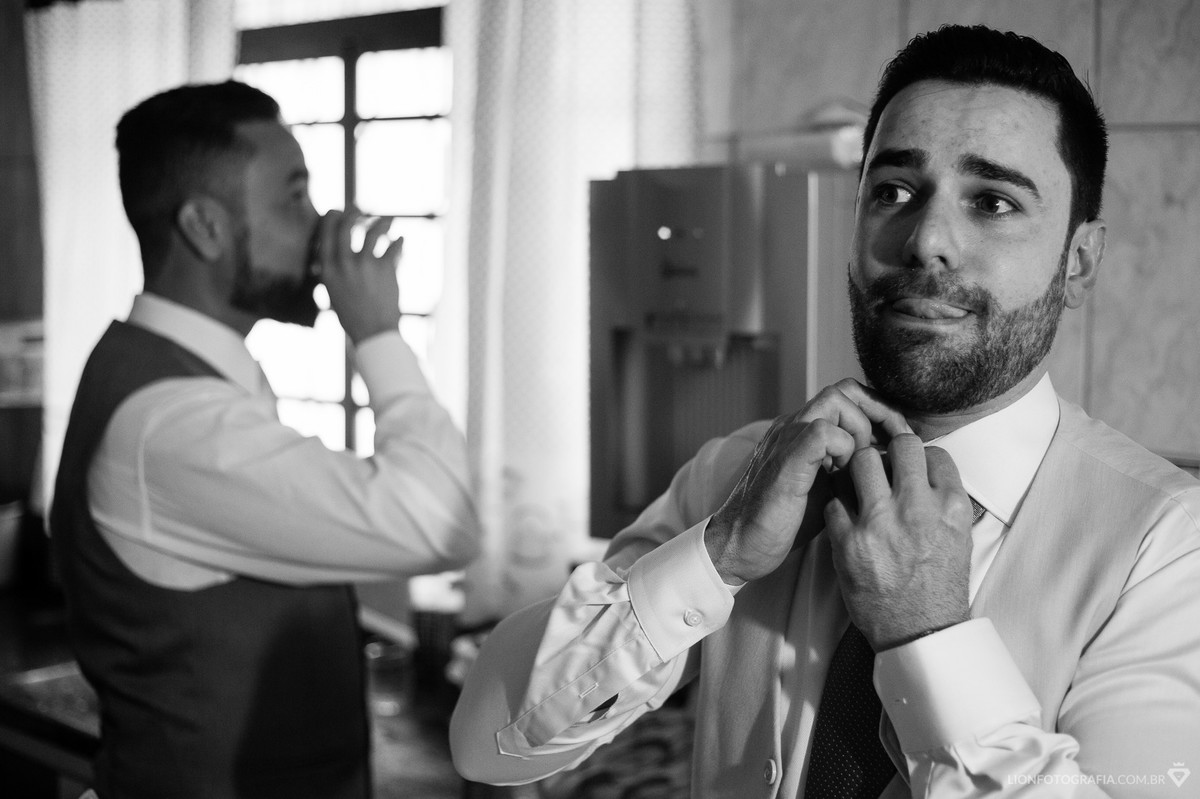 O noivo Armando apertando o nó da gravata enquanto seu amigo e padrinho bebe água antes do casamento