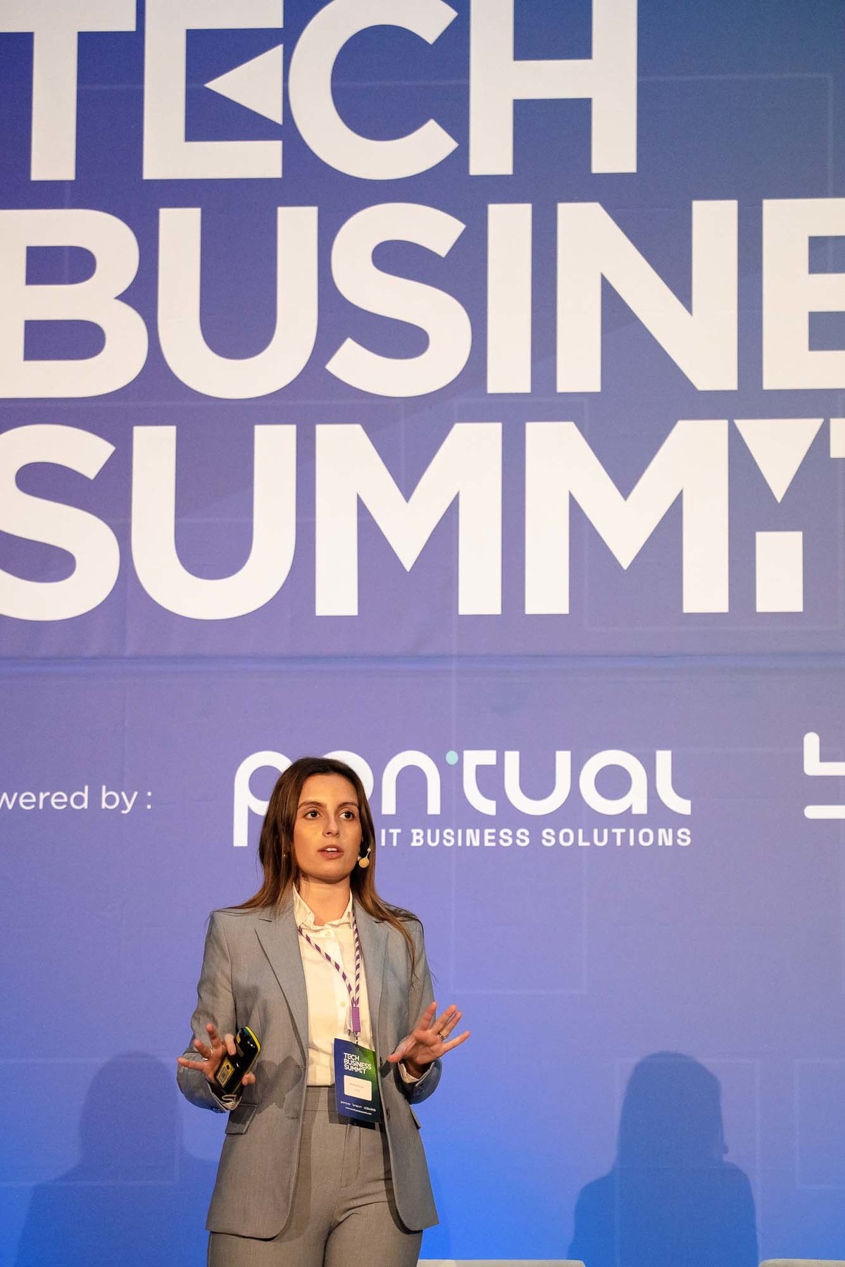 Orador em pé no palco com microfone na mão, a apresentar com entusiasmo.       