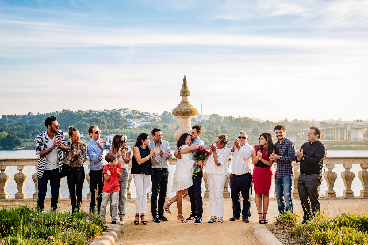 Imagem vibrante dos noivos ao centro e os amigos e familiares em fila, todos a celebrar a boa notícia com alegria