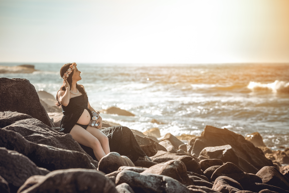 gestante sentada nas pedras com o mar em um belo céu azul ao fundo e usando vestido de renda azul marinho