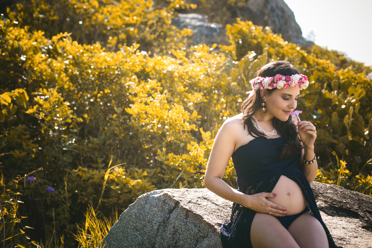 Ensaio de Gestante  sentada na pedra com  a mão na barriga e cheirando uma flor