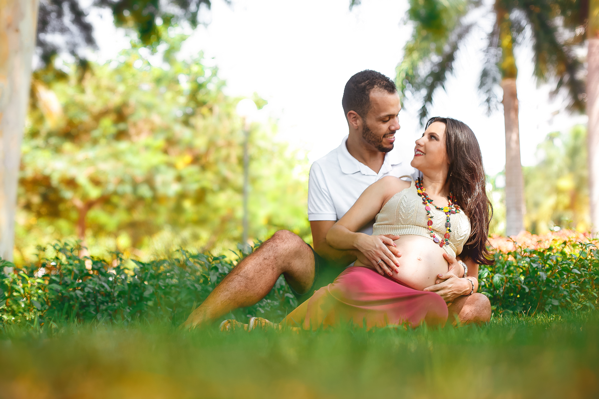 Ensaio de gestante ela olha  pro esposo sentada na grama