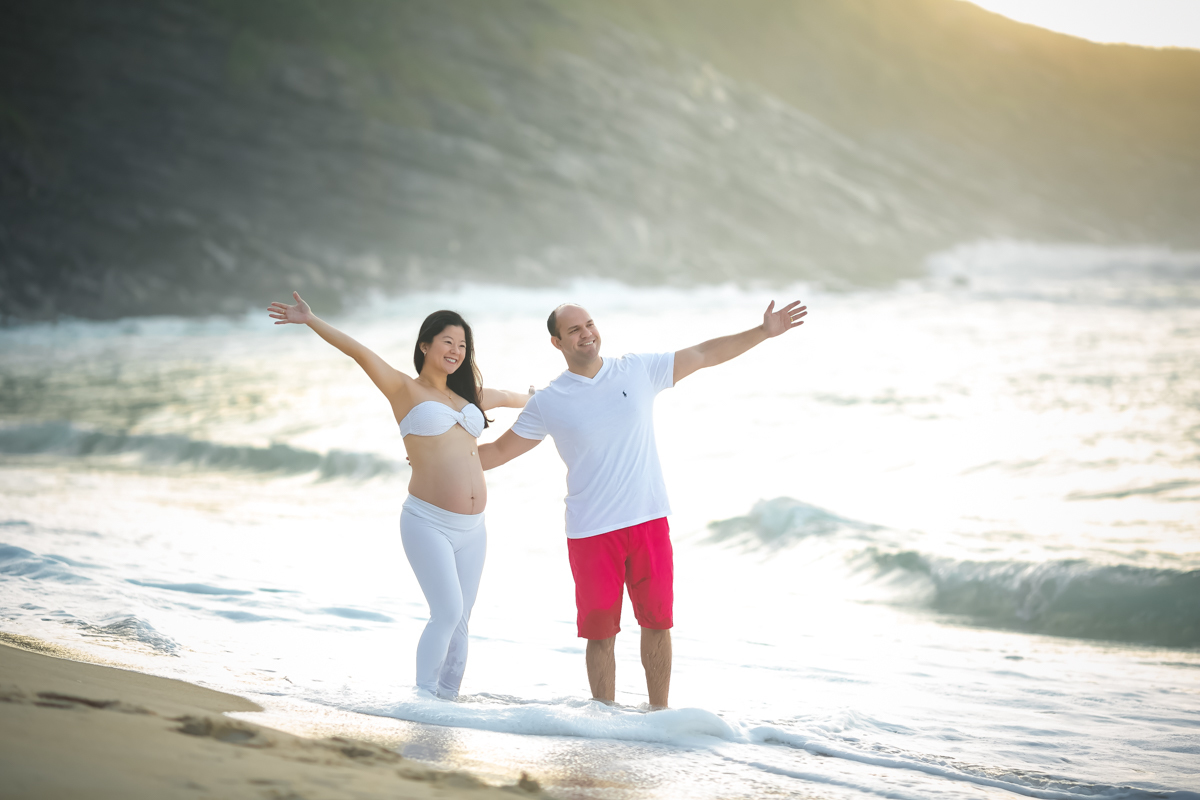 Ensaio externo de  gestante junto com esposo com os braços abertos caminhando na praia de Grumari-RJ