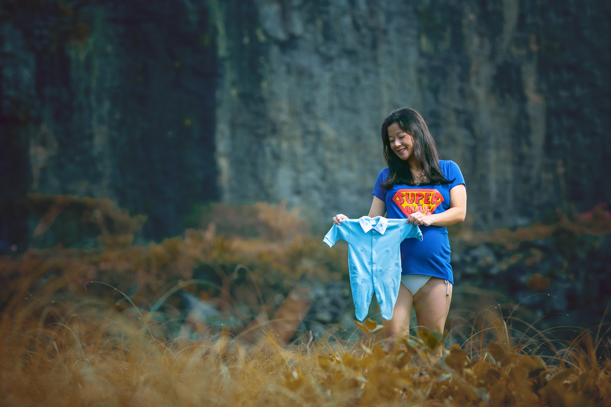 Book gestante na praia segurando roupinha, e no fundo paredão de  pedras na Prainha-RJ 