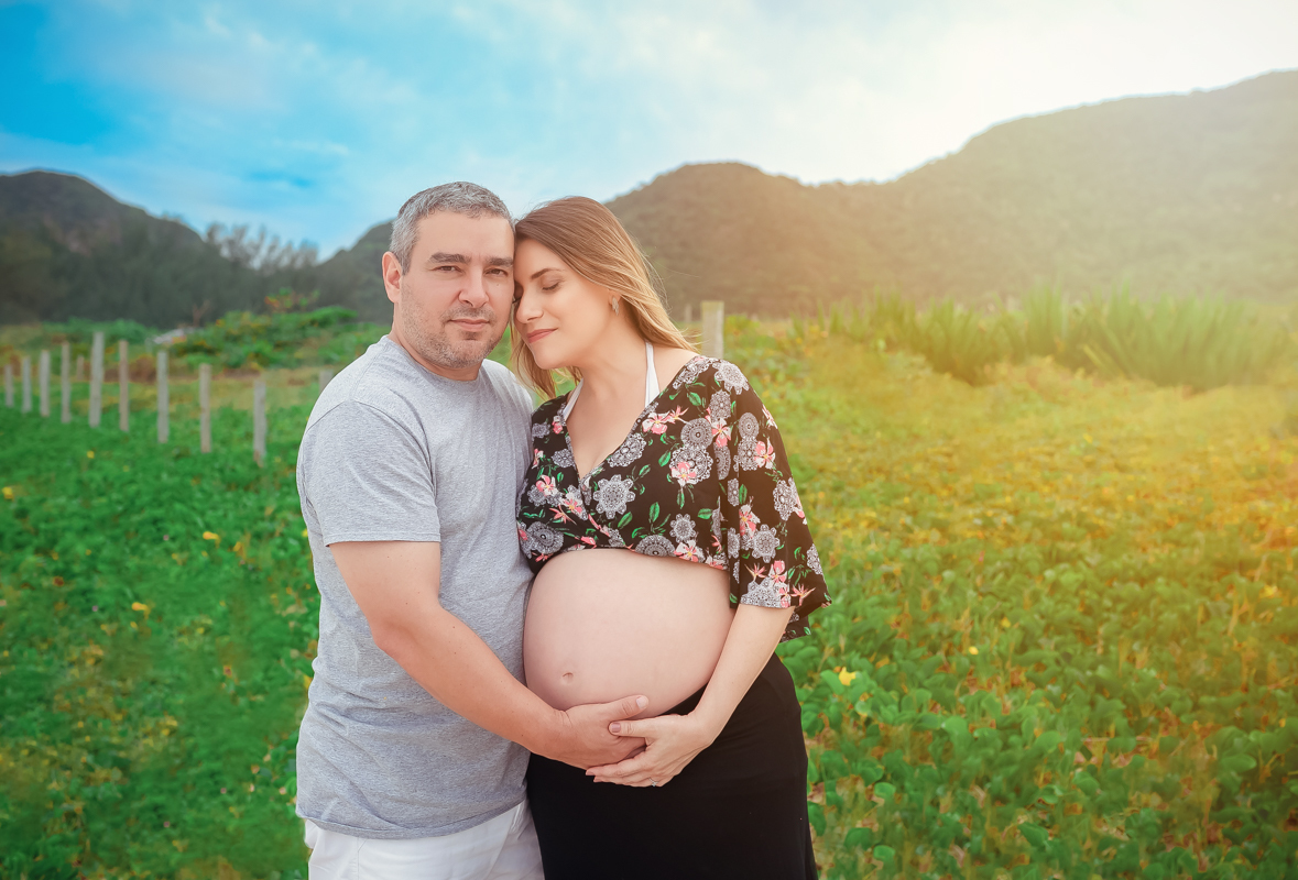 Casal em ensaio fotográfico de gestante esposo colocando mão em sua barriga/Grumari