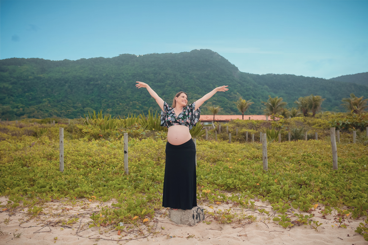 Gestante de braços abertos ao fundo muito verde em seu ensaio externo em Grumari/RJ
