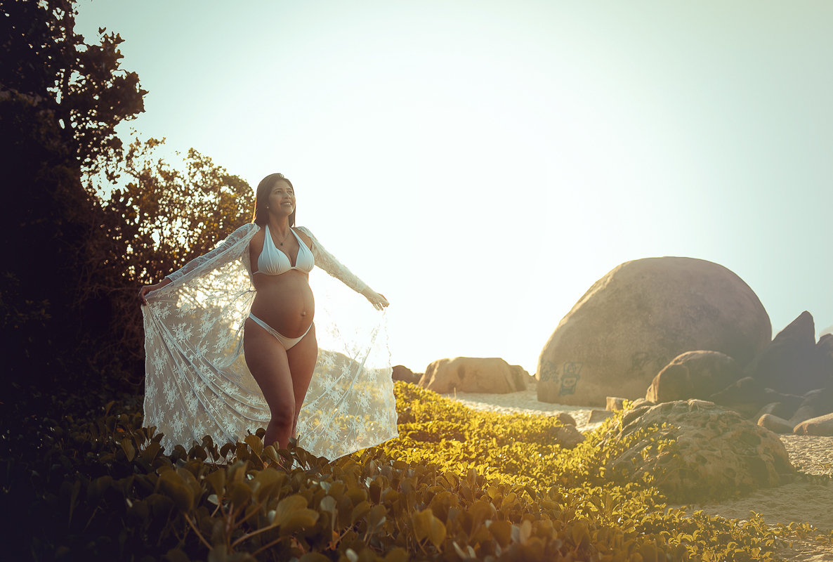 Gestante com os braços abertos contemplando a natureza em ensaio na prainha