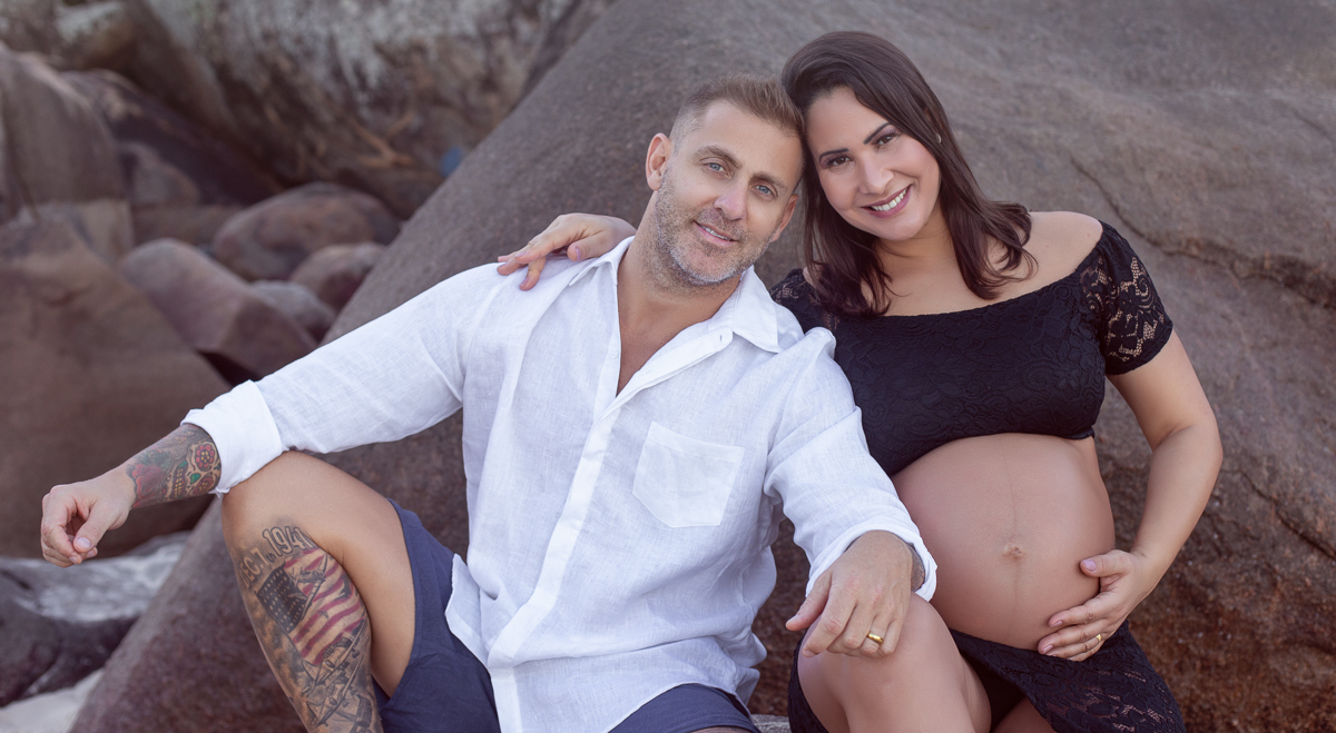 gestante sentada na pedra posando para foto junto com seu esposo em ensaio fotográfico em praia/rj