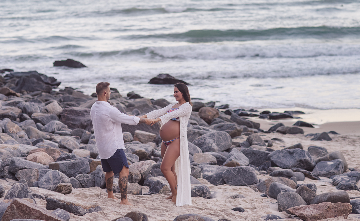 gestante de mãos dadas com seu esposo e ao fundo mar e pedras em ensaio fotográfico