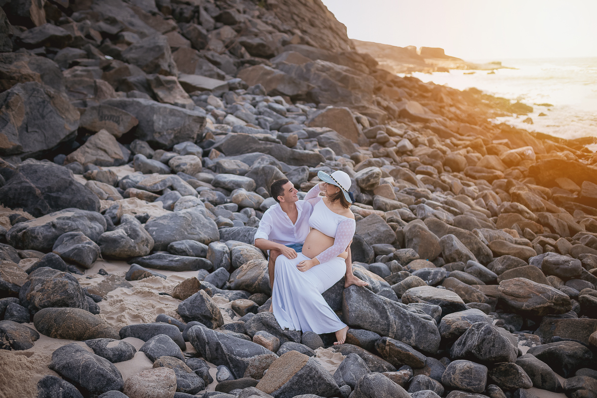 gestante sentada no colo de seu esposo ao fundo pedras mar-prainha-RJ