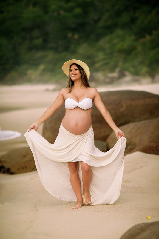gestante caminhando na praia , segurando saia fazendo pose em seu ensaio fotográfico externo