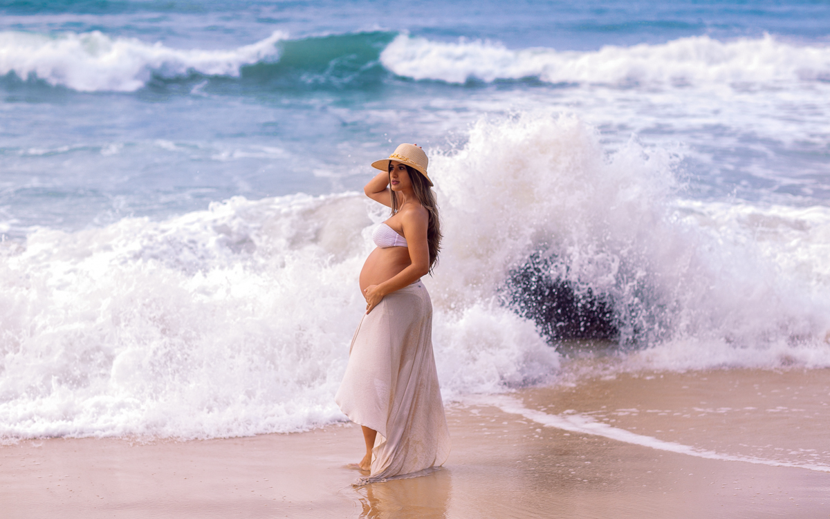 ondas de quebrando ao fundo, e gestante fazendo pose em seu ensaio fotográfico extreno