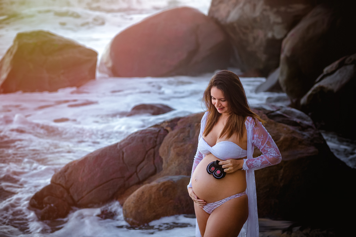 gestante fazendo pose apoiando sapatinho na barriga ao fundo pedras em seu ensaio fotográfico externo