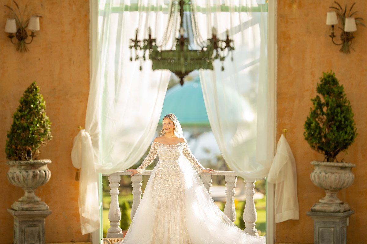 Ainda na frente do pallazo nossa linda Manú, está de frente no centro da fotografia colorida, com as mãos apoiadas na sacada com a luz do sol radiante batendo em seu véu.