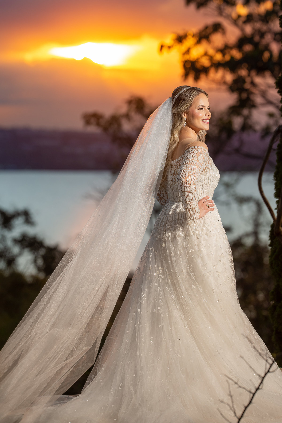 Mais um lindo retrato colorido dessa noiva linda, com o finalzinho do por do sol o céu em degradê, a noiva está meio de costas, com as mãos na cintura, com seu véu para trás e um lindo sorriso no rosto.