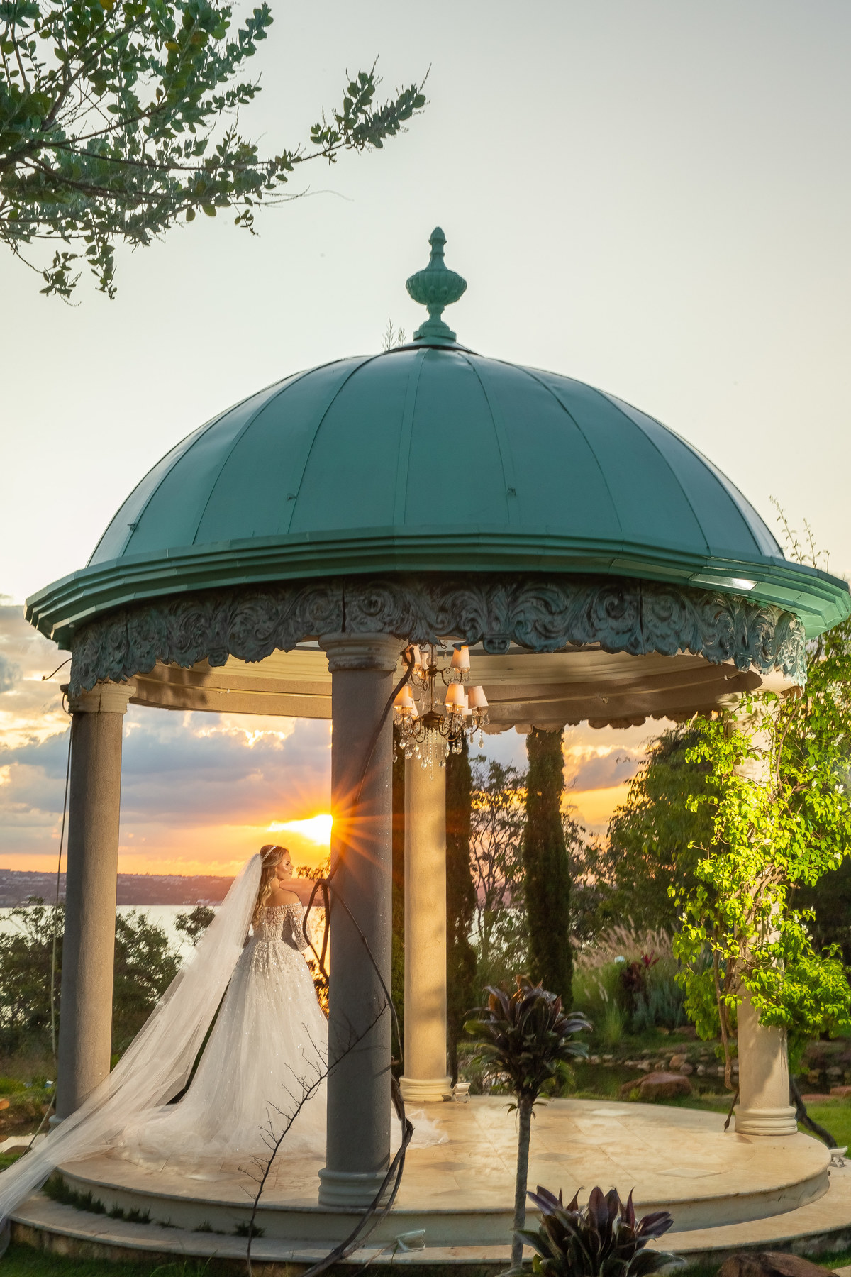 Uma foto maravilhosa ainda no por do sol, a noiva dentro do gazebo, os raios do sol entram lateral a ela formando um flair,  o céu está lindo com muitas nuvens, tudo perfeito. 