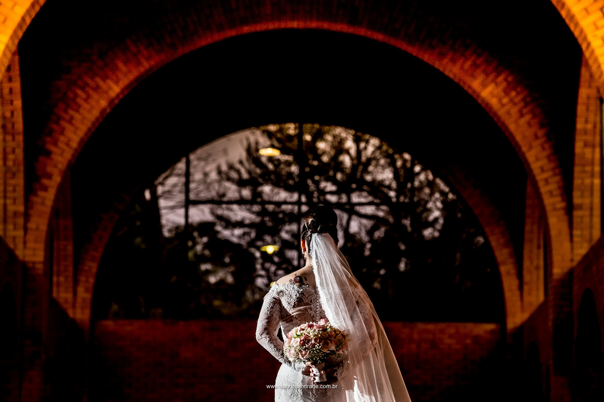 Nesta fotografia colorida o fotografo Sandro Andrade pode brincar com sua maravilhosa técnica de luz e fez uma fotografia criativa da noiva de costas evidenciando seu buque e uma luz belíssima do por do sol, entre os arcos no céu.