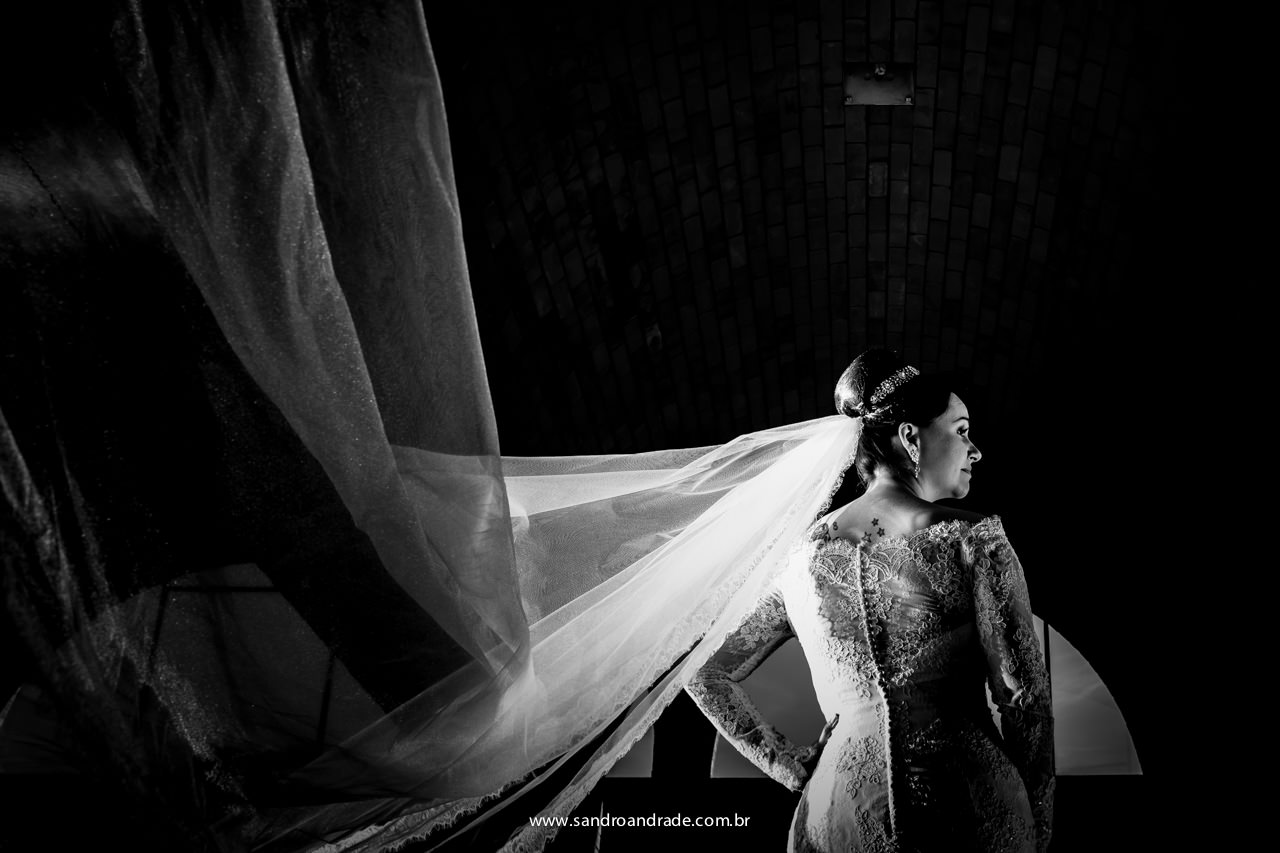 Uma linda fotografia preto e branco, mostrando em detalhes as costas de seu vestido de noiva e seu véu ao vento fazendo um contorno lindo contorno compondo esta fotografia.