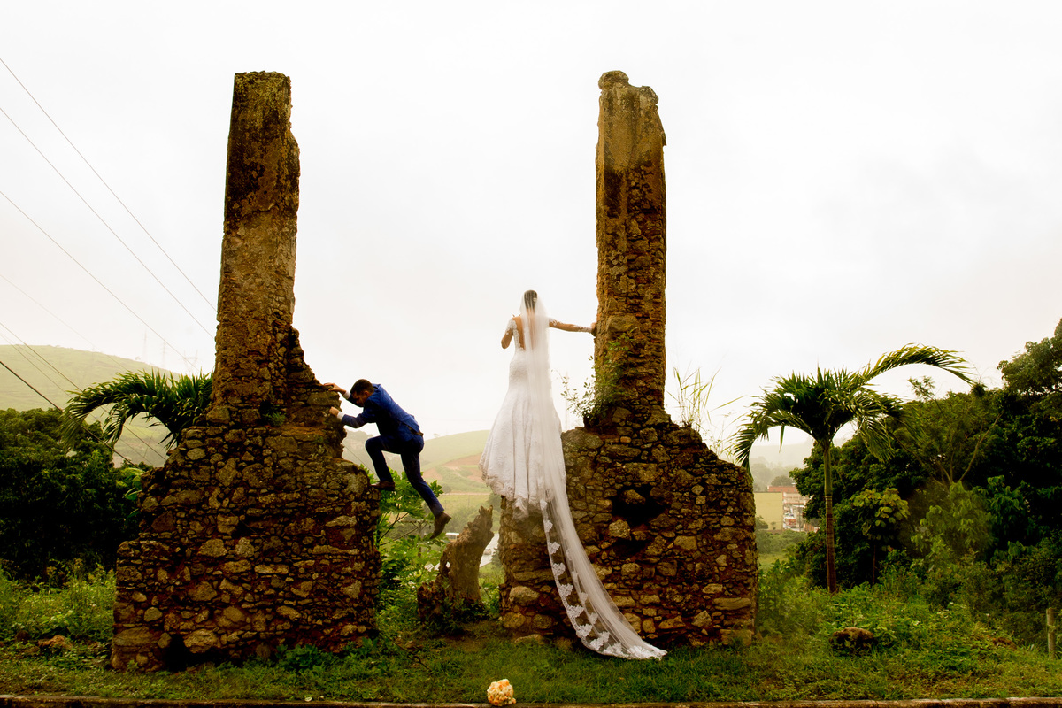 A noiva em cima de um muro em ruínas e seu noivo subindo na outra ponta, fotografia espontanea. 