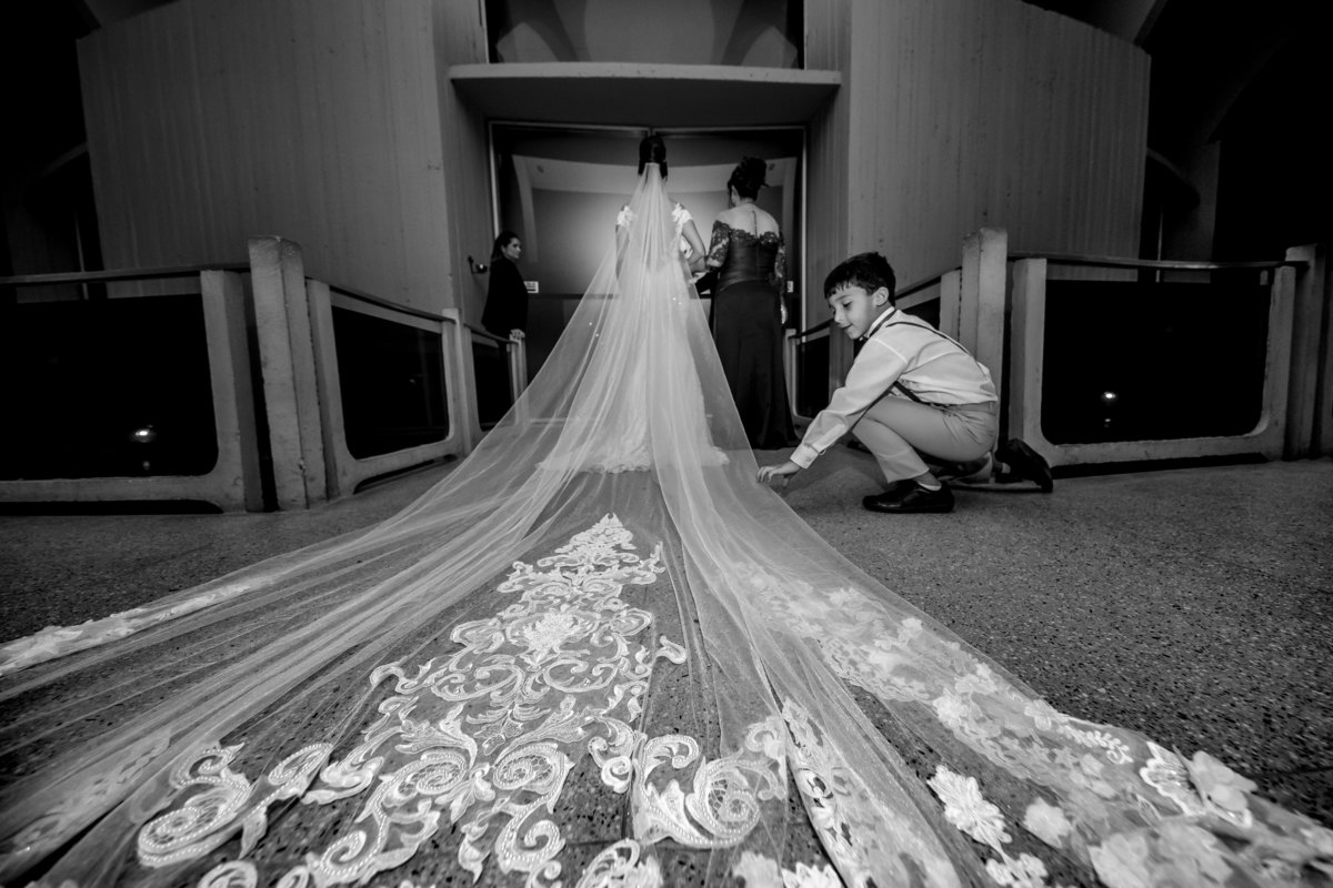 Na porta da igreja de braço dado com a mãe, Priscila aguarda seu momento de entrar, enquanto isso o pajem que levará as alianças arruma com carinho véu da noiva, sua tia.