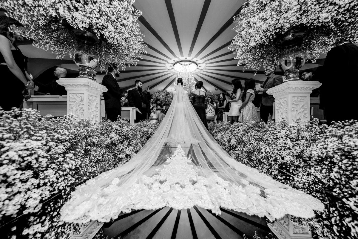 Um detalhe do véu da noiva, em uma fotografia preto e branco e toda a belíssima decoração compondo esta fotografia da entrada da noiva, feita de costas.