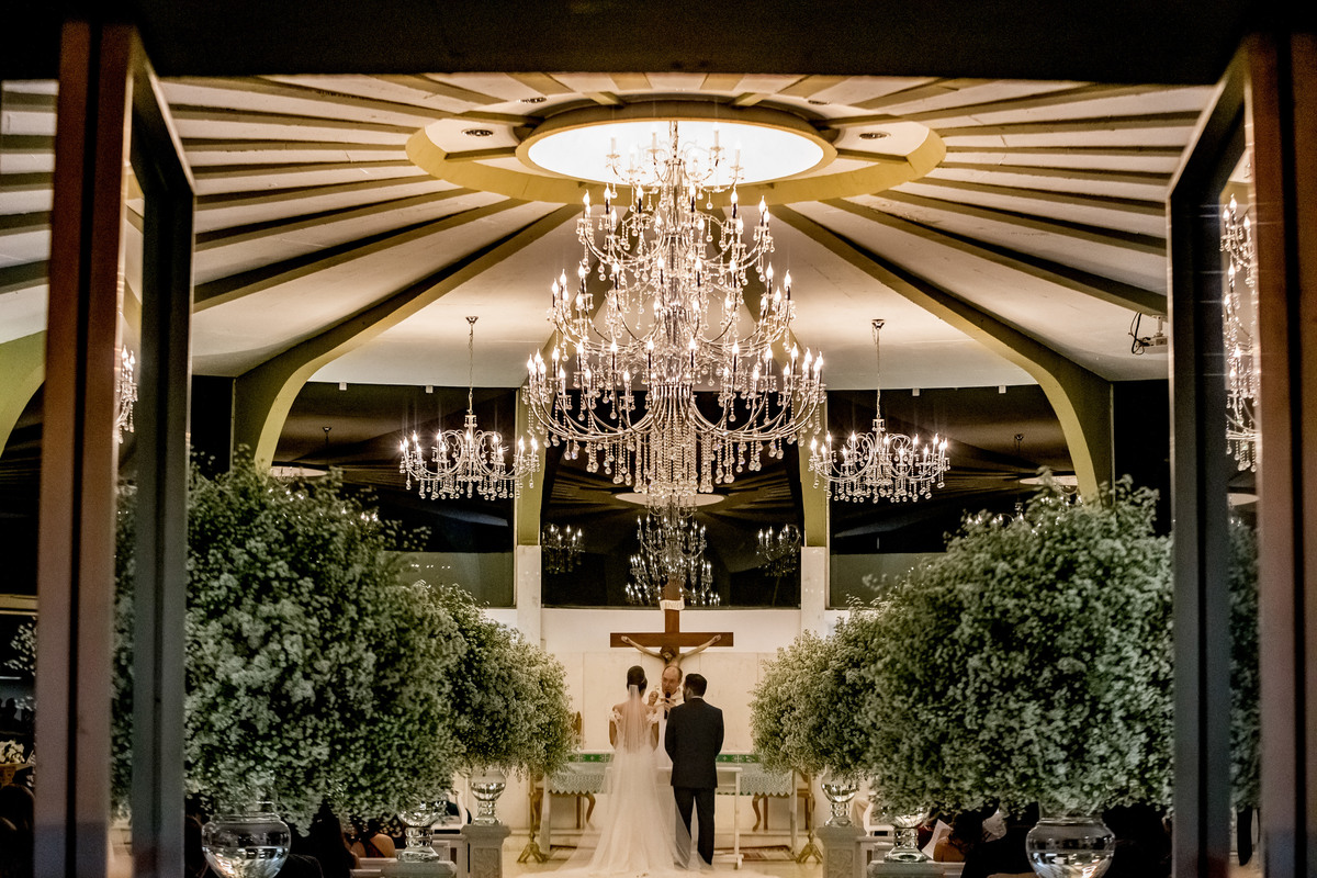 Como já é de costume nos casamentos Sandro Andrade nosso fotografo premiado e talentosíssimo, sempre procura uma fotografia nova, um olhar diferente para cada casal, assim é esta foto, o casal no altar entre as portas, mostrando os lustres e a cruz.