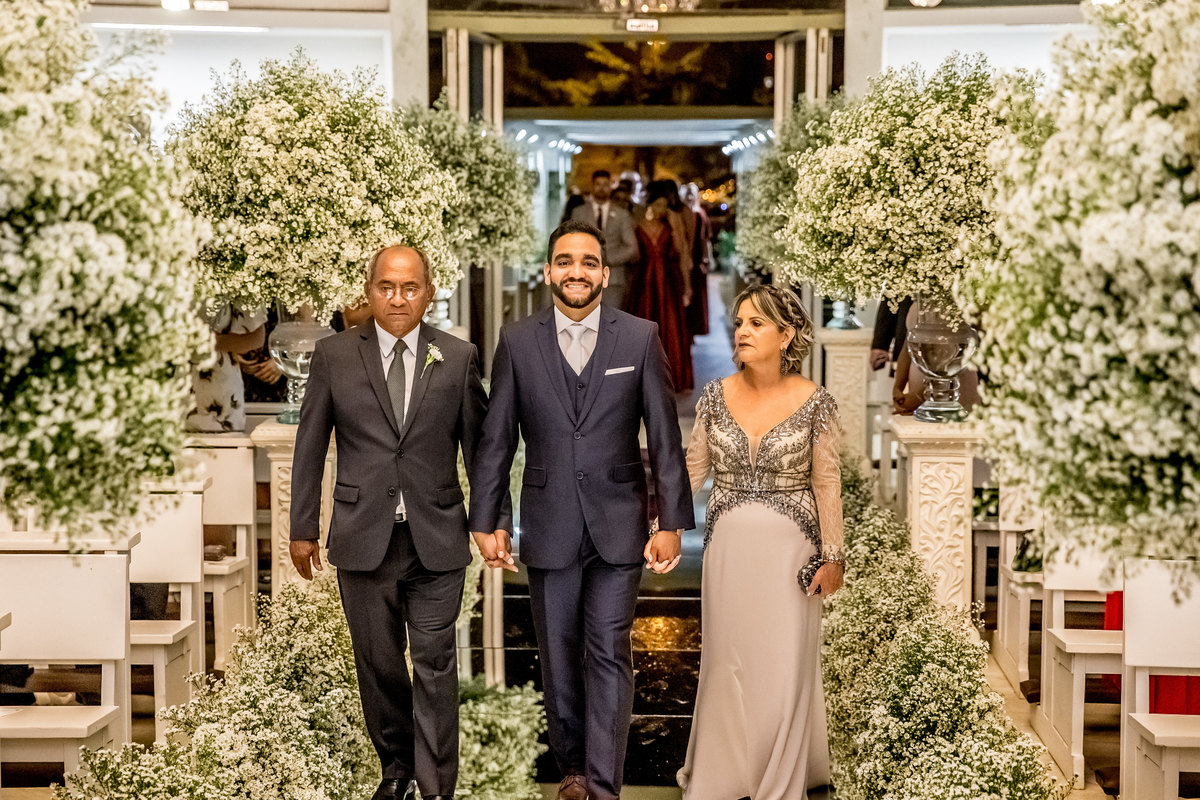 A entrada do noivo em uma linda fotografia colorida, ele entra com a igreja bem decorada por Kaká Fagundes, de mãos dadas com os pais e todos alegres e sorridentes.