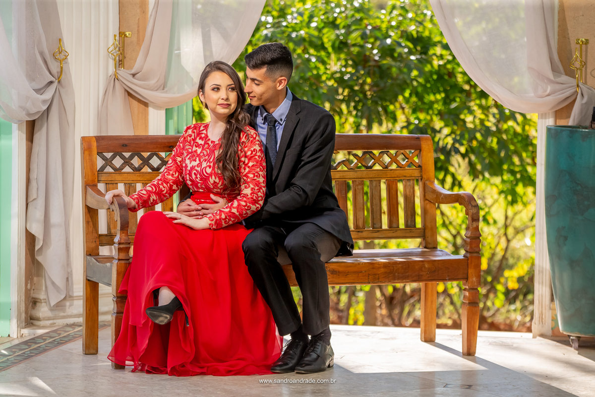 Retrato dos noivos, sentados no banco de madeira o noivo admira sua noiva, Tons bem quentes complementam esta fotografia, o sol ao fundo, a noiva de vestido vermelho e o noivo de terno preto com uma camisa azul.