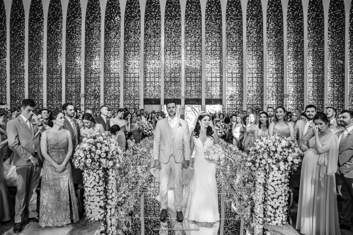 Uma fotografia preto e branca, com os noivos no centro, a arquitetura da igreja e dos convidados nas laterais, mostrando a emoção de cada um.