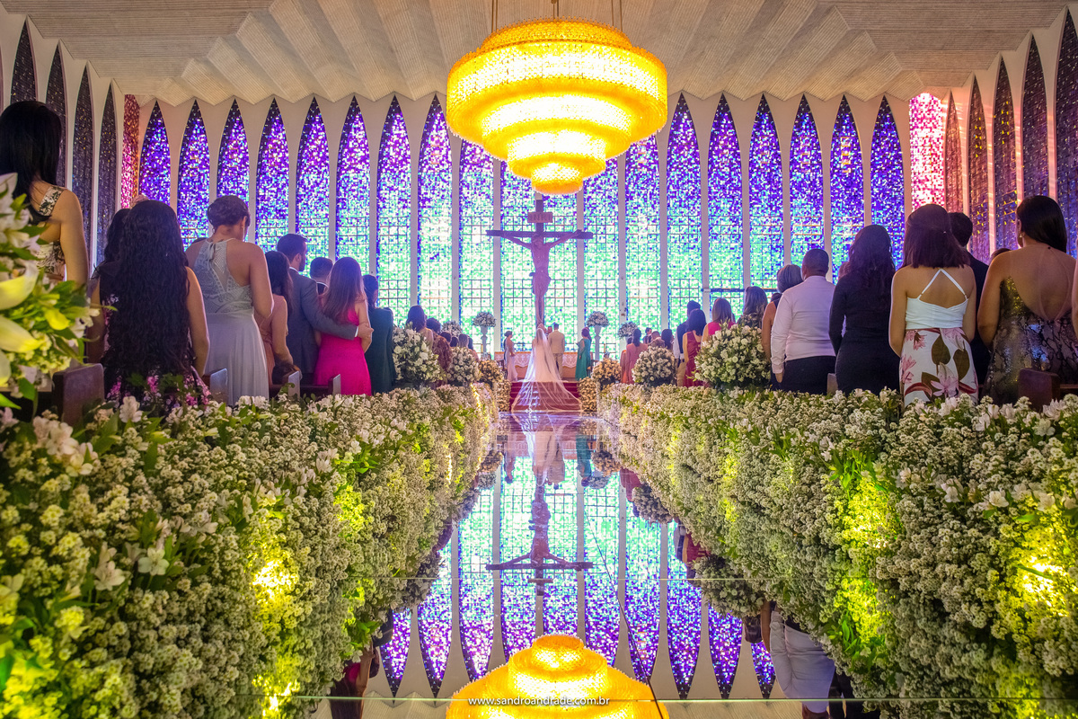 Um registro perfeito do casal recebendo a benção inicial, fotografia pegando a igreja toda de costas, com todo o altar e a decoração de flores e tapete espelhado, refletindo o teto, o luz magnifico e o casal com seus pais e padre.