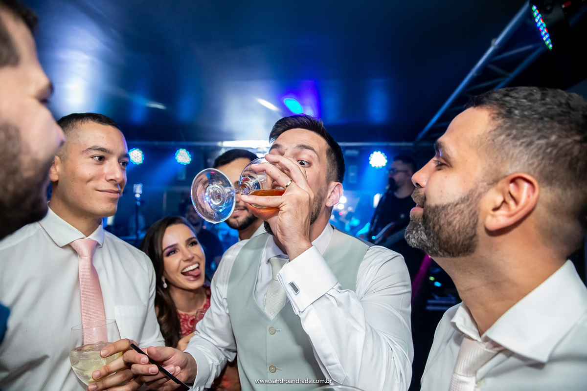 Os amigos do noivo observam ele virar uma taça de drink do open bar.