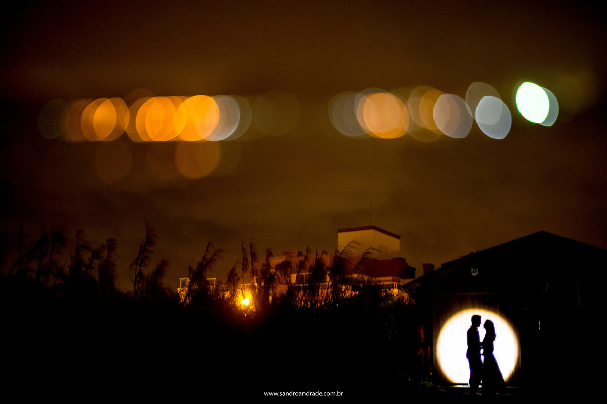 Obra de arte, no breu da praia, o fotógrafo de casamentos de brasilia, Sandro Andrade visualiza uma foto e parte em ação, a cabana serve de moldura para o casal que com uma contra luz na mão faz um circulo e a silhueta do casal está centro dele e bonquês.