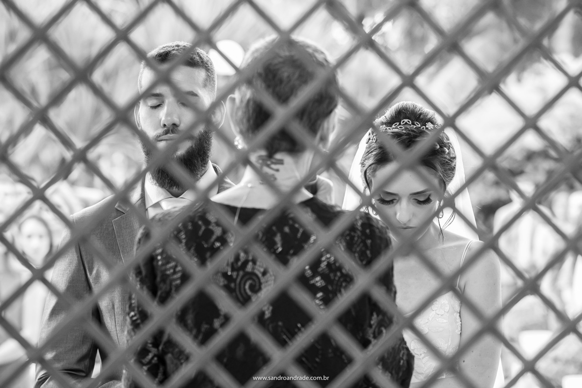 Entre as grades do pergolado, a celebrante de costas e no meio dos noivos, ainda orando de olhos fechado nas laterais.