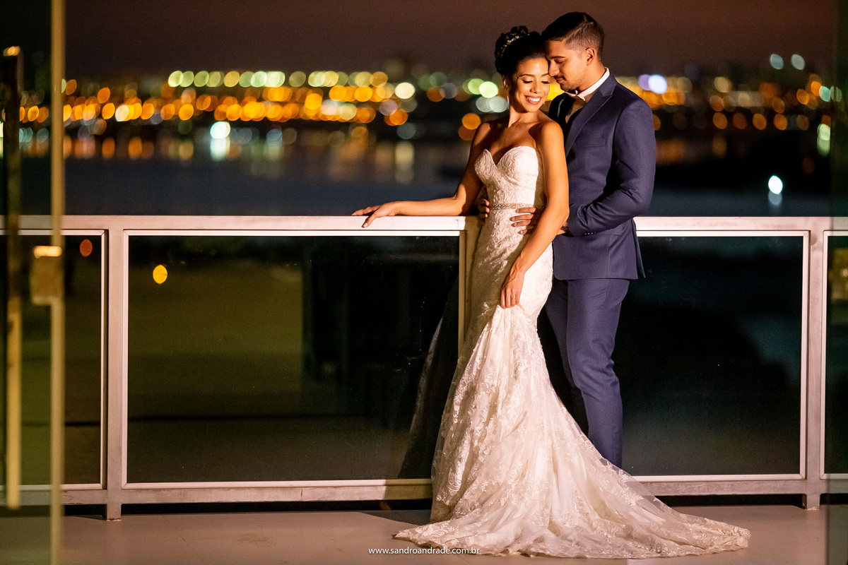 De volta a varanda, os noivos estão abraçados e toda a vista do lago com as bolinhas de luz do outro lado aparecendo, linda fotografia feita por um dos melhores fotógrafos de Brasilia.