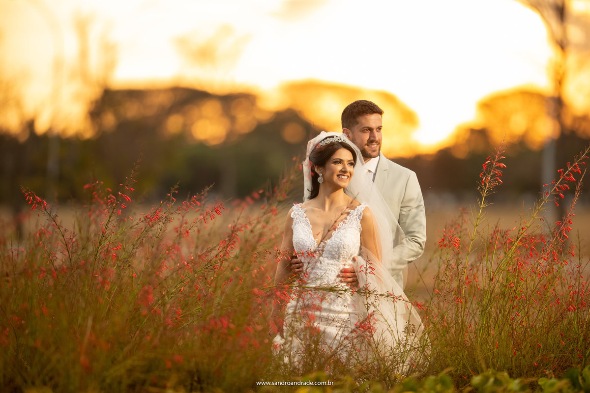 No meio de pequeninas flores vermelhas, o casal está ao centro desta imagem, sorrindo, Luiza de costas para Michell e um belo por do sol ao fundo.